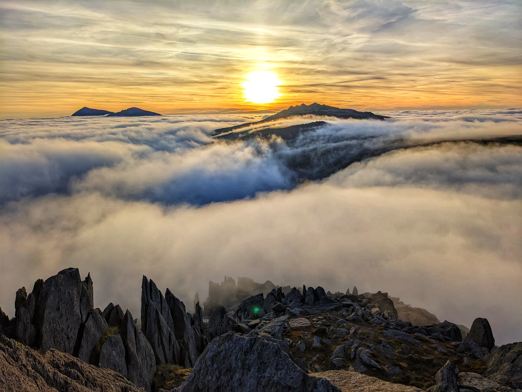 Sunlit mountain summits above a sea of cloud, with jagged rocks in the foreground