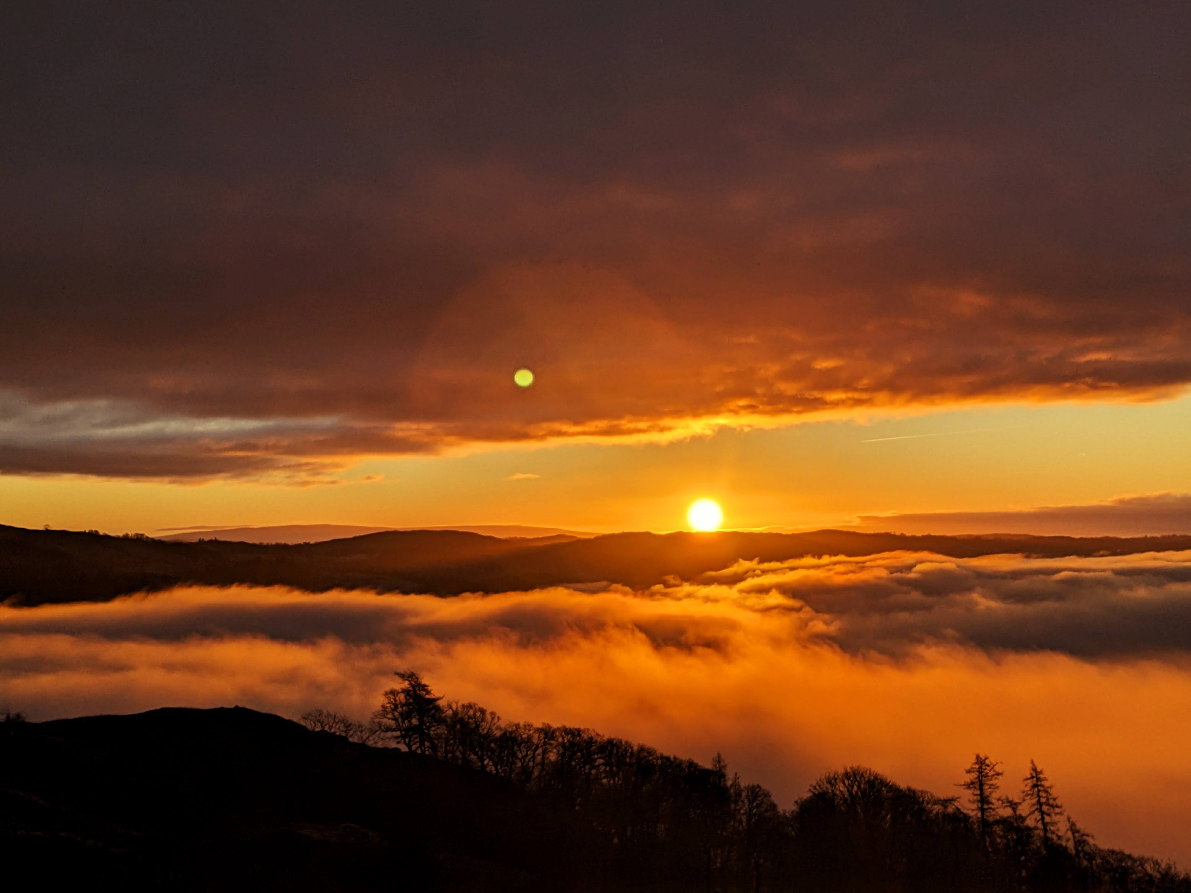Golden sunrise above low cloud filling a valley, with silhouetted hills and trees in the foreground